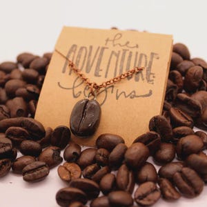 Può includere: Una collana con ciondolo a forma di chicco di caffè con una catena marrone, esposta su un letto di chicchi di caffè. Un piccolo cartoncino marrone dietro la collana recita "the ADVENTURE begins". Il ciondolo è un chicco di caffè marrone scuro.