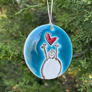 May include: A round ceramic ornament with a glossy turquoise blue glaze featuring a hand-painted white snowman with brown outlines reaching up toward a bright red heart. The ornament hangs from a thin, light-colored string loop at the top.