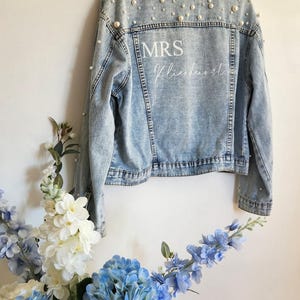 May include: A light blue denim jacket decorated with faux pearls, with the text "MRS Klienhorst" printed on the back. The jacket is displayed against a wall, with a floral arrangement.