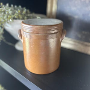 May include: A brown stoneware crock with a lid. The cylindrical container has two small handles and horizontal ridges near the top. The lid is a lighter shade of brown. The crock is approximately 6 inches tall and is a vintage kitchen decor item.