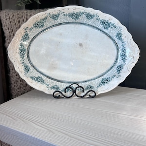 May include: An antique oval serving platter with a white center and a teal floral border. The edges are scalloped and the platter is supported by a black metal stand. A decorative piece for serving or display.