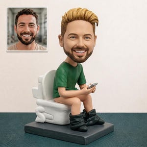 May include: A custom bobblehead figurine featuring a person with light brown hair and a beard, wearing a green t-shirt and black trousers pulled down around the ankles, sitting on a white toilet while holding a small grey smartphone in their hands.
