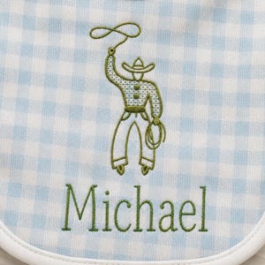 May include: Baby bib with a light blue and white gingham pattern. The bib features an embroidered green cowboy design and the name "Michael" in green lettering. The bib has a white trim.