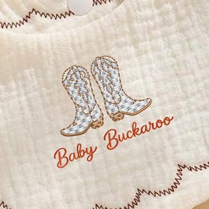 May include: Cream-colored baby bib with embroidered cowboy boots and the text "Baby Buckaroo" in a reddish-brown color. The bib has a scalloped edge with brown stitching and a snap closure.