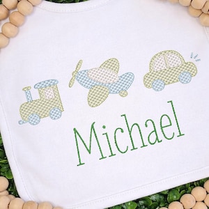 May include: White baby bib with embroidered train, airplane, and car in green and blue. The name "Michael" is embroidered in green script below the vehicles. The bib is surrounded by a wooden bead garland.