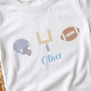 May include: White t-shirt with embroidered football-themed design. Features a blue football helmet, a gold goalpost, and a brown football. The name "Oliver" is embroidered in blue script below the goalpost.