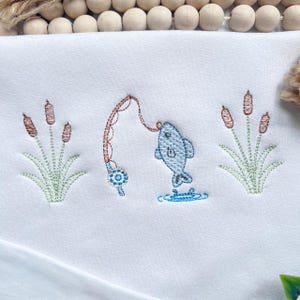 May include: A white fabric surface featuring an embroidered design of a fishing rod catching a blue fish, flanked by two clusters of brown cattails with green stems and leaves. The embroidery uses blue, brown, and green thread on white fabric.
