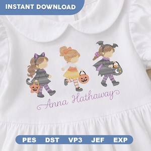 May include: A white shirt featuring an embroidered design of three children in Halloween costumes, including a cat, a pumpkin, and a bat. The text Anna Hathaway is embroidered in purple script below the figures. The shirt has a rounded collar.