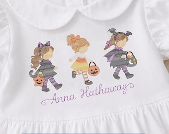 May include: A white shirt featuring an embroidered design of three children in Halloween costumes, including a cat, a pumpkin, and a bat. The text Anna Hathaway is embroidered in purple script below the figures. The shirt has a rounded collar.