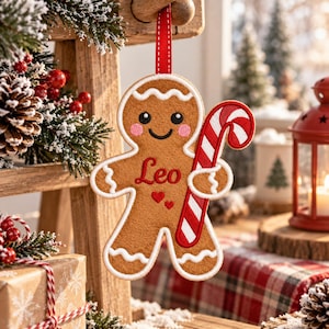 May include: A tan felt gingerbread man Christmas ornament with white icing trim, pink cheeks, and a red and white candy cane. The name Leo is embroidered in red script on the chest above two small red hearts, suspended by a red ribbon.
