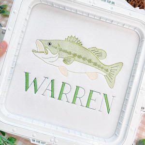 May include: A white fabric square secured in a white plastic embroidery hoop, featuring an embroidered green and light-colored bass fish above the name WARREN stitched in green capital letters with a dark green outline on the white fabric.