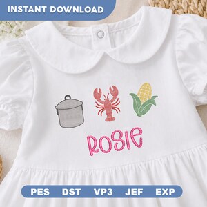 May include: A white short-sleeved dress featuring an embroidered design of a grey cooking pot, a red lobster, and a yellow ear of corn. Below the embroidery, the name Rosie is stitched in pink cursive lettering. The dress has a rounded collar.