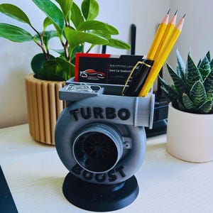 Peut inclure: Un organiseur de bureau gris et noir en forme de turbocompresseur, avec les mots "TURBO BOOST" sur le devant. L'organisateur contient des crayons, un stylo, des cartes de visite et des trombones. Une petite plante en pot est visible en arri&egrave;re-plan.