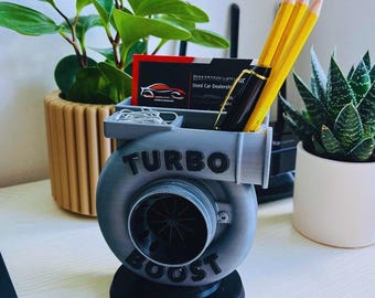 May include: A gray and black desk organizer shaped like a turbocharger, with the words "TURBO BOOST" on the front. The organizer holds pencils, a pen, business cards, and paper clips. A small potted plant is visible in the background.