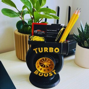 Peut inclure: Organisateur de bureau noir et or en forme de turbocompresseur, avec les mots "TURBO BOOST" en lettres dor&eacute;es. Il contient des crayons, un stylo, des cartes de visite et des trombones jaunes. Une petite plante et une succulente sont en arri&egrave;re-plan.