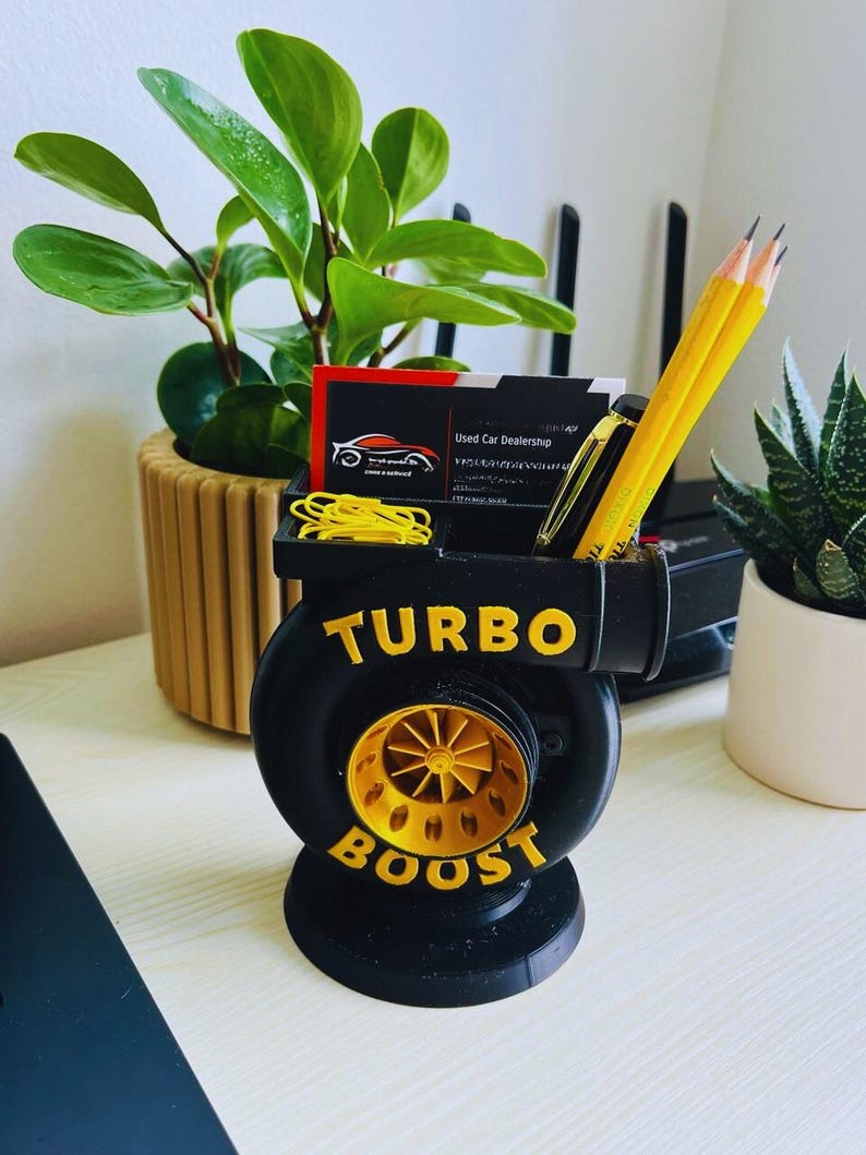 Peut inclure: Organisateur de bureau noir et or en forme de turbocompresseur, avec les mots "TURBO BOOST" en lettres dor&eacute;es. Il contient des crayons, un stylo, des cartes de visite et des trombones jaunes. Une petite plante et une succulente sont en arri&egrave;re-plan.
