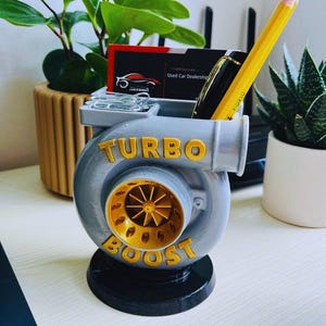 Peut inclure: Un organiseur de bureau gris et dor&eacute; en forme de turbocompresseur, avec les mots "TURBO BOOST" en lettres dor&eacute;es. Il contient un crayon, un stylo, des cartes de visite et des trombones. Des plantes vertes sont en arri&egrave;re-plan.