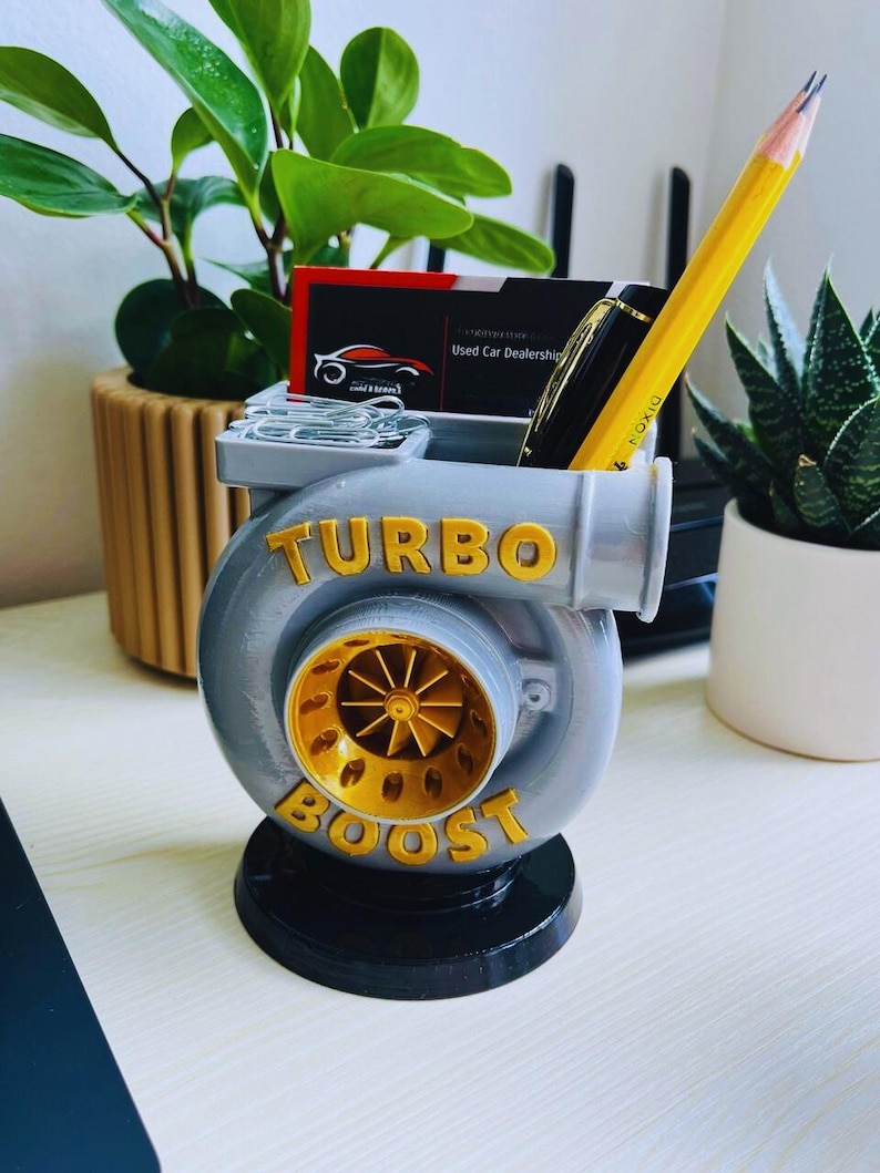 Peut inclure: Un organiseur de bureau gris et dor&eacute; en forme de turbocompresseur, avec les mots "TURBO BOOST" en lettres dor&eacute;es. Il contient un crayon, un stylo, des cartes de visite et des trombones. Des plantes vertes sont en arri&egrave;re-plan.