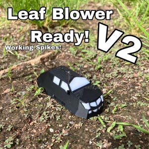 Könnte beinhalten: Ein schwarzes Miniaturfahrzeug mit weißen Akzenten und zwei Antennen, beschriftet mit "Leaf Blower Ready!" und "Working Spikes!" mit einer "V2" Grafik. Das Spielzeug steht auf einem Beet aus Erde und kleinen Pflanzen.