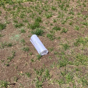 May include: A rolled-up white paper with the word "Shelter" printed on it, lying on the ground. The ground is covered with patches of grass and dirt. The image is taken outdoors in natural light.