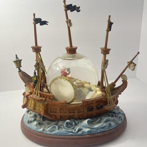 May include: A decorative ship-shaped snow globe featuring a brown wooden-style hull, three masts with black flags, and two small lanterns. The central clear glass sphere contains a small figure, and the base is sculpted to resemble blue ocean waves.