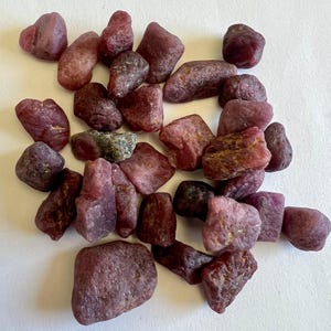 Könnte beinhalten: Eine Ansammlung roher Rubin-Edelsteine in verschiedenen Formen und Größen. Die Steine zeigen eine Reihe von tiefroten und rosa Farbtönen, wobei einige eine dunklere, fast violette Tönung aufweisen. Die raue Textur der Edelsteine ist deutlich sichtbar.