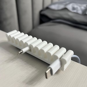 May include: A white cable tidy with multiple slots, holding two charging cables with different connector types. The organiser is on a light-coloured surface, with a blurred background.