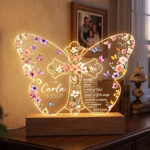May include: A butterfly-shaped LED night light with floral and butterfly accents. The light includes a cross design and the name "Carla" with the date "05.02.2026". The light sits on a wooden base.