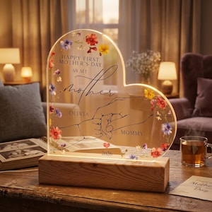 Può includere: Targa in acrilico a forma di cuore con decorazioni floreali e base in legno. La targa recita "Happy First Mother's Day as my Mother" con i nomi "Olivia" e "Mommy" e l'anno "2026". Il design presenta due mani che si tengono.