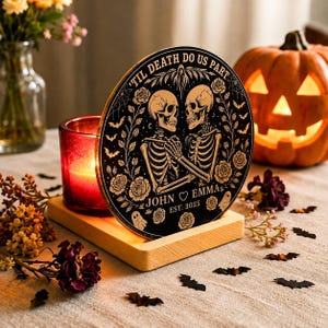 Op de afbeelding: Een ronde zwarte plaat met een goudkleurige illustratie van twee skeletten die elkaars hand vasthouden, omringd door rozen, vleermuizen en spinnenwebben. De tekst luidt 'TIL DEATH DO US PART', 'JOHN HEART EMMA' en 'EST. 3025'. Het staat op een lichte houten voet naast een rode kaars.
