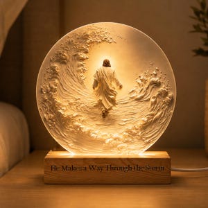 May include: A circular, illuminated acrylic lamp featuring a relief sculpture of a figure walking through turbulent waves. The light glows from behind the figure. The lamp sits on a rectangular wooden base engraved with the text, He Makes a Way Through the Storm.