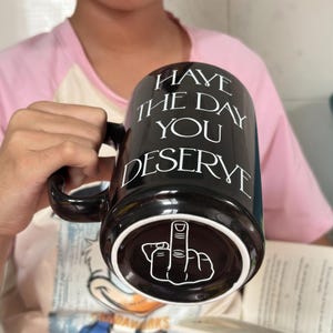 Może przedstawiać: Czarny ceramiczny kubek z białym napisem "HAVE THE DAY YOU DESERVE" nadrukowanym z przodu. Spód kubka przedstawia białą grafikę liniową dłoni wykonującej gest środkowego palca, widoczną pod kątem od dołu naczynia.
