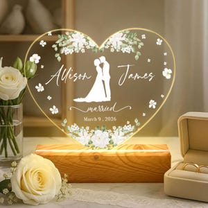 Könnte beinhalten: Eine herzförmige Acrylplatte mit Holzsockel, die eine weiße Silhouette eines Paares, florale Akzente sowie den Text Allison, James, married und March 9, 2026 zeigt. Die Platte ist beleuchtet und mit weißen Blüten und grünen Blättern verziert.