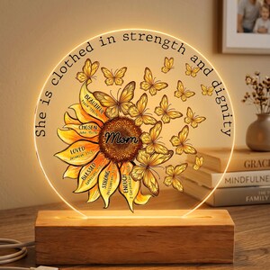 May include: An LED lamp with a sunflower and butterfly design. The clear acrylic disc has the words "She is clothed in strength and dignity" and other inspirational words. The lamp sits on a wooden base.