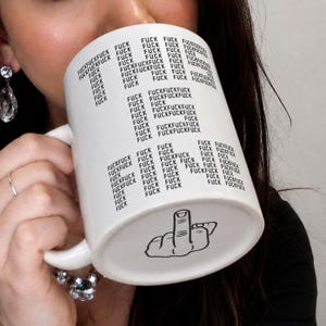 May include: A white ceramic mug featuring the word FUCK printed repeatedly in black text across the surface, arranged in columns. At the bottom of the mug, there is a black line drawing of a hand making a middle finger gesture.