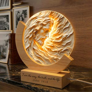 Può includere: Una lampada circolare con una scultura in rilievo di una figura barbuta con capelli fluenti, inserita in una cornice di legno chiaro. La base è incisa con il testo: I am the way, the truth, and the life. John 14:6.