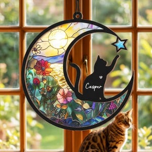 Puede incluir: Adorno de ventana redondo de vidriera con una silueta de gato negro que alcanza una estrella azul. El gato está posado en una luna creciente con el nombre "Casper". El fondo representa un paisaje colorido con flores y un sol.