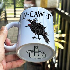 May include: A white ceramic mug featuring a black and white illustration of a crow with its beak open and the text F-CAW-F printed above it. The base of the mug displays a black and white graphic of a hand making a middle finger gesture.