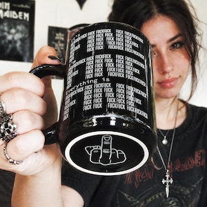 Può includere: Una tazza in ceramica nera con il testo bianco ripetuto "It's everything is FUCK" che copre la superficie esterna. La base della tazza mostra un'illustrazione grafica bianca di una mano che fa il gesto del dito medio su sfondo nero.