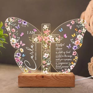 May include: A butterfly-shaped acrylic night light with a wooden base. The design features a cross and floral accents, with the name "Carla" and the date "10.12.2023". Text includes "I AM", "beautiful", "chosen", "a child of God", "created in His image", "made for a purpose", "a conqueror", and "loved forever".