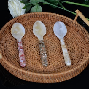 Puede incluir: Tres cucharas decorativas con cuencos de nácar y mangos ornamentados en tonos rojos, plateados y crema, presentadas en una bandeja de mimbre trenzado con asas de madera. Las cucharas están dispuestas en fila.