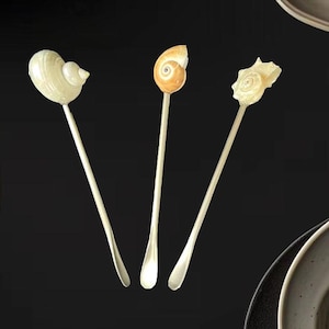 Puede incluir: Tres cucharas decorativas con tapas en forma de concha. Las cucharas tienen mangos largos y delgados y están dispuestas sobre un fondo oscuro. Las conchas son blancas y naranjas.