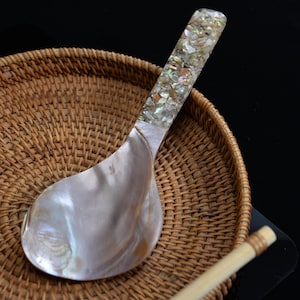 Puede incluir: Una cuchara de servir nacarada con un mango con incrustaciones de fragmentos de concha iridiscente. La cuchara descansa sobre una bandeja de mimbre tejido, con un fondo negro. El cuenco de la cuchara es de un blanco opalescente brillante.