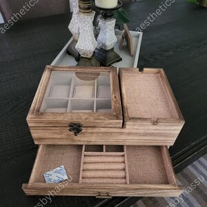 May include: A wooden jewelry box with a glass top and multiple compartments. The box has a drawer with ring slots and a separate tray. The interior is lined with a beige fabric. The box is on a dark wooden table.