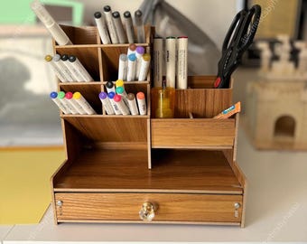 May include: A wooden desk organizer with multiple compartments for pens, markers, and scissors. The organizer has a drawer with a crystal knob. The wood grain finish gives it a natural look. Various colored markers and pens are visible.