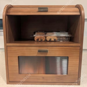 May include: A wooden bread box with a roll-top lid and a lower drawer with a ribbed glass panel. The box is made of light-colored wood and has a simple, modern design. Inside, there are pastries in a clear plastic container.