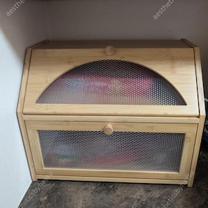 May include: A light-colored bamboo bread box with a mesh front and a curved, mesh-covered window on the top. The box has a small wooden knob on the door and top. The bread box is sitting on a dark surface.