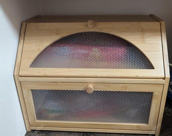May include: A light-colored bamboo bread box with a mesh front and a curved, mesh-covered window on the top. The box has a small wooden knob on the door and top. The bread box is sitting on a dark surface.