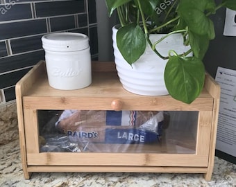 May include: A bamboo bread box with a clear glass door, a white ceramic butter dish, and a white potted plant. The bread box contains a loaf of bread. The word "LARGE" is visible on the bread packaging.