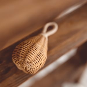 Könnte beinhalten: Eine gewebte Rassel aus Rattan mit Schlaufengriff. Die Rassel hat eine tropfenförmige Form und besteht aus hellbraunem, gewebtem Material. Der Griff ist heller und scheint aus einer weichen Naturfaser zu bestehen. Die Rassel liegt auf einer Holzoberfläche.